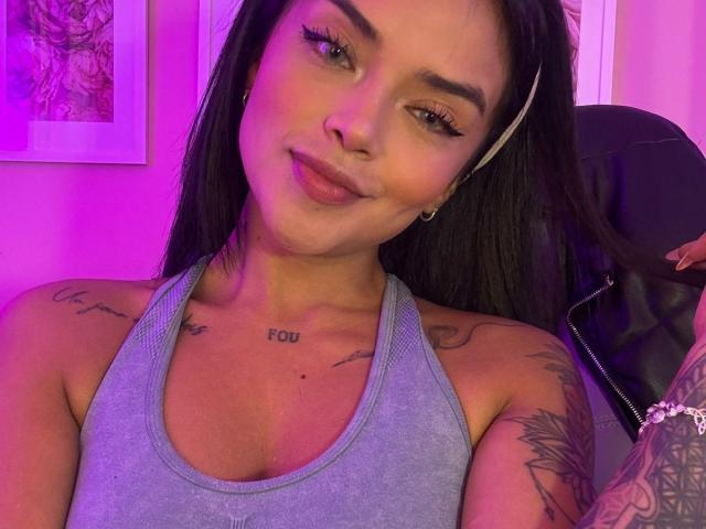 MajoJones with medium tits, tattoos, and a gray tank top, smiling at the camera, purple lighting, black hair, and a white headband, showing a confident and sexy look.