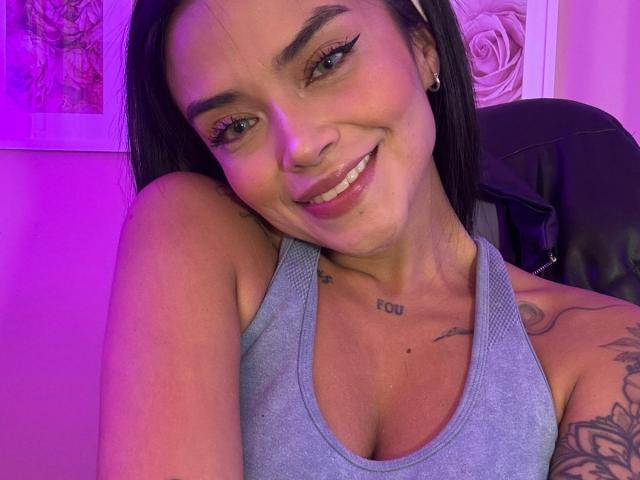 MajoJones with black hair, blue eyes, and a smile, wearing a grey tank top, showing tattoos on her chest and arm, and a nose ring, under pink lighting.