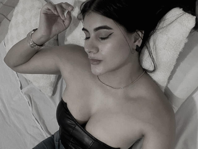 AlaraKey is sleeping with her eyes closed, wearing a strapless black leather top that reveals her large boobs. She has dark hair, a chain necklace, and a bracelet.