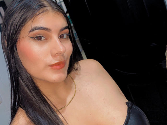 AlaraKey with long black hair, eyeliner, and orange lipstick, wearing a black strap top and gold necklace, showing her tits and a subtle nose piercing.