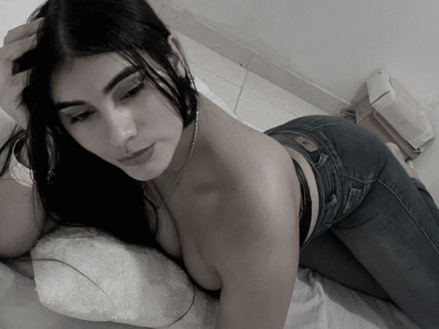 AlaraKey with long black hair, lying topless on a bed, wearing blue jeans, showing her ass, looking at the camera with a slight smile, hand in her hair.
