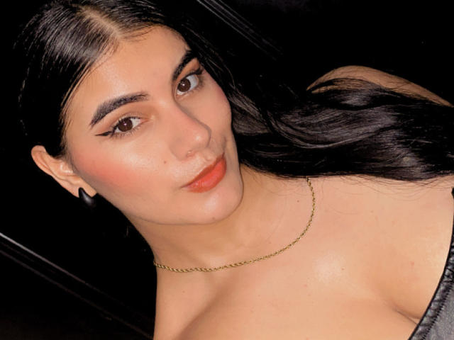 AlaraKey has dark, wavy hair, brown eyes, and red lipstick. She's wearing a gold chain necklace and has a slightly upturned nose with a small, subtle nose ring.