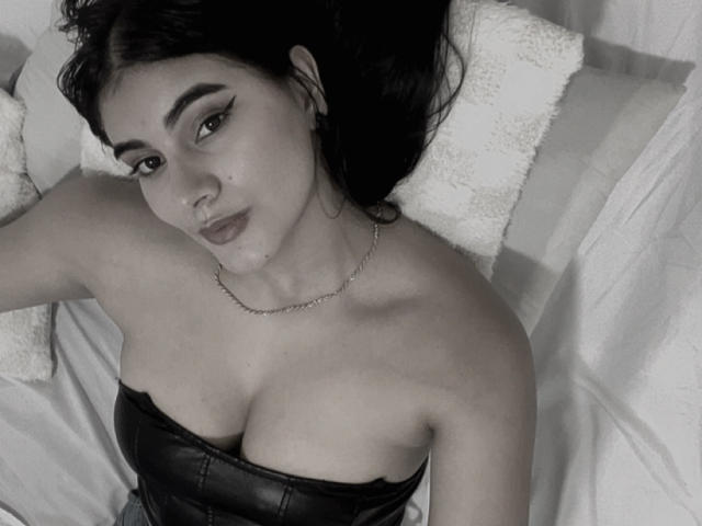 AlaraKey with dark hair, thick eyebrows, and eyeliner, lying on bed, wearing a strapless black leather top, showing her big tits, and a chain necklace.
