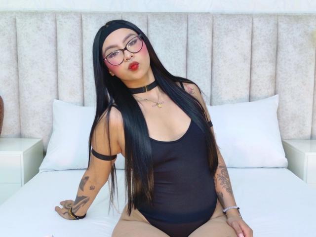 ChloeMoretti with long black hair, glasses, red lipstick, black choker, black tank top, showing tattoos, sitting on a bed with white pillows and a beige headboard.