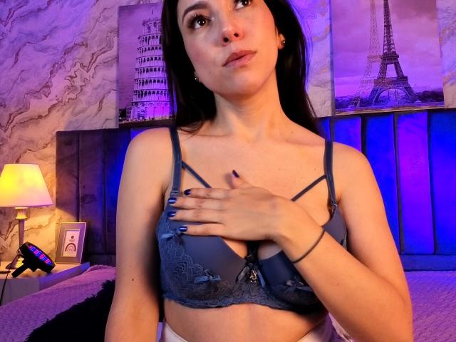 MiaReed with blue lace bra, hand on chest, dark hair, purple-blue lighting, looking up, bedroom, purple and blue tones, black nail polish, serious expression, bra covering her tits.