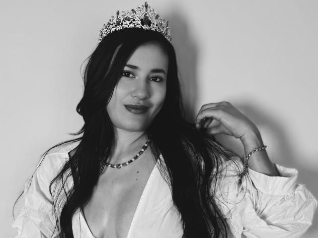 SarahMoonOne with long black hair, wearing a jeweled crown, white shirt, deep cleavage, beaded necklace, and bracelet, smiling slightly, touching her hair. Black-and-white photo.