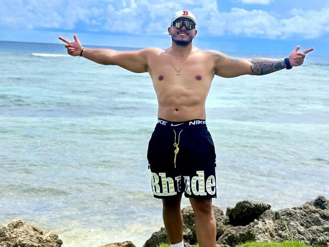 OwenPierce, shirtless, standing on rocks, arms out, wearing black Nike shorts, "Rhude" text, white cap, sunglasses, tattooed arm, necklace, and pendant, pointing with fingers.