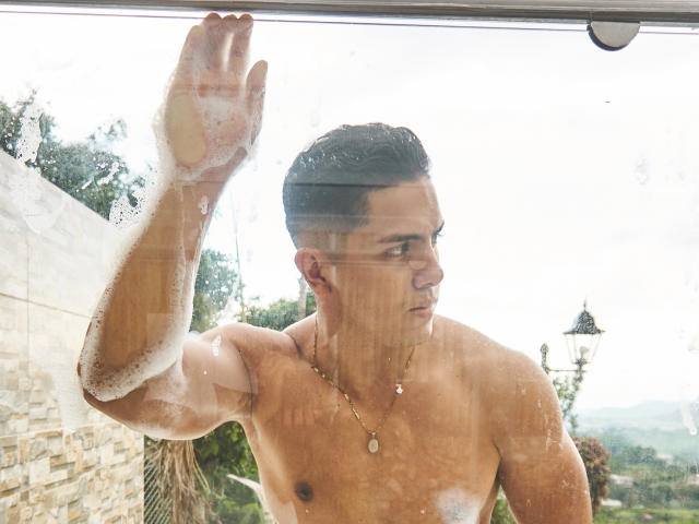 OwenPierce, shirtless, soap suds on arm, raising hand against glass, looking to the side, short dark hair, necklace with pendant, muscular, wet skin, outside, daytime, slightly foggy glass.