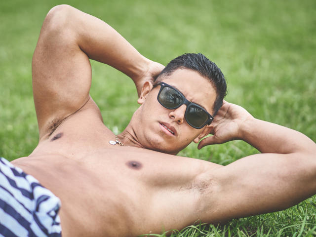 OwenPierce, shirtless, wearing black sunglasses, lying on grass with arms behind head, showing armpit hair, striped shorts, and a small necklace, looking serious.