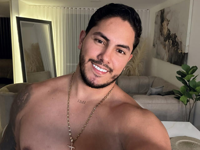 OwenPierce, shirtless, smiling, short dark hair, beard, tan skin, gold necklace, chest tattoo, toned muscles, brown eyes, looking at camera, living room, plant in background.