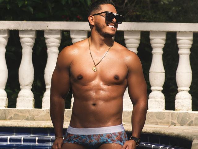 OwenPierce has a muscular, sweaty, brown-skinned body, wearing blue patterned briefs, black sunglasses, and a gold necklace, sitting by a tiled pool with a white balustrade.