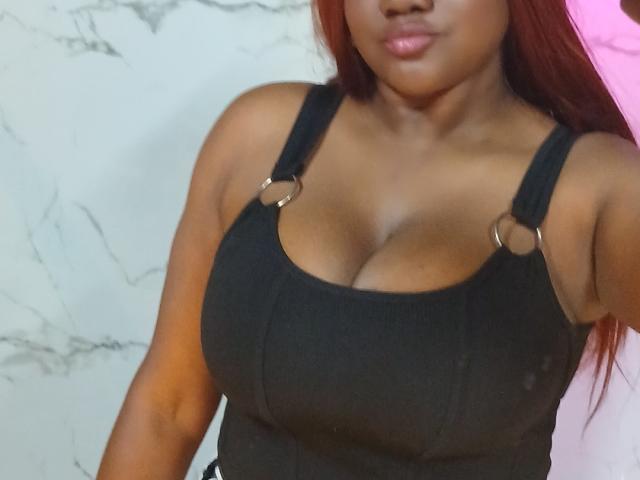 RoseBeckers with dark skin, red hair, wearing a black tank top with metal rings, showing large, round tits and a pouty face, in a slightly blurry image.