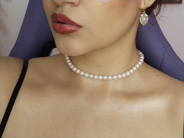 JesicaBomShell's face, showing glossy red lips, a pearl necklace, and gold heart-shaped earring, with a visible black strap on her shoulder. Her skin is shiny and tan.