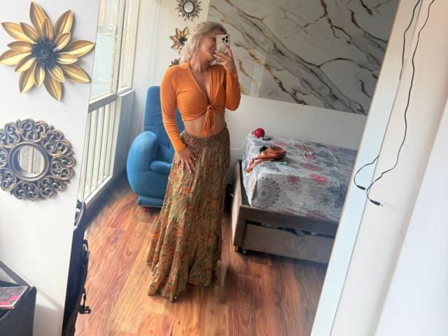 FloraSaenz with curly blonde hair, orange long-sleeve top, and patterned skirt, standing in bedroom, holding phone, mirror selfie, blue chair, wooden floor, marble wall.