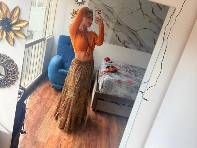 FloraSaenz with orange crop top, tied at the front, showing her small tits, and a long, flowy, patterned skirt, taking a selfie in a mirror, standing on wooden floor.
