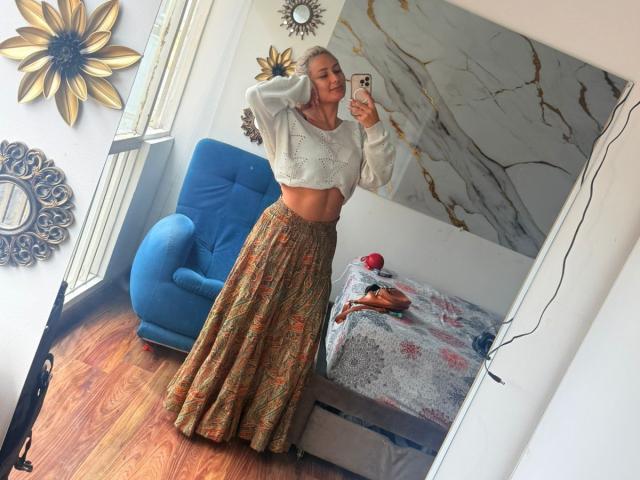 FloraSaenz stands in a mirror selfie, wearing a white crop top and a long, patterned skirt, showing her toned midriff and blonde hair, with a blue chair nearby.
