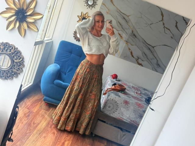 FloraSaenz with blonde hair, white crop top, and colorful skirt, holding phone, standing in room, blue chair, marble wall art, wooden floor, mirror selfie, smiling.