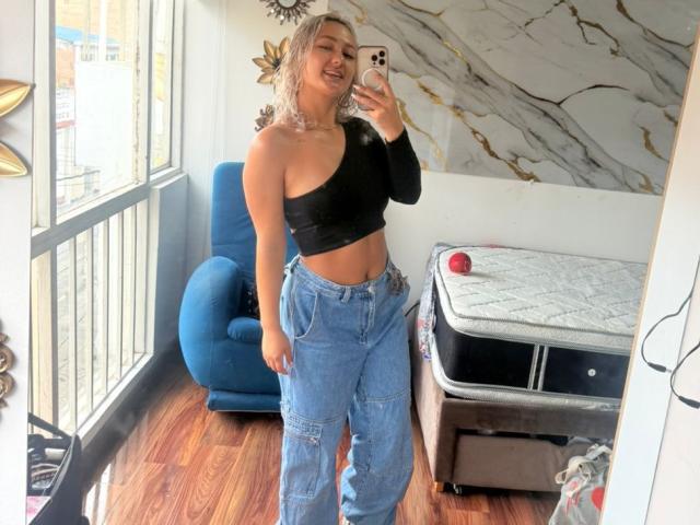 FloraSaenz with blonde hair, black one-shoulder top, and blue jeans, smiling in a mirror selfie, showing her toned midriff and ass, standing on wooden floors.