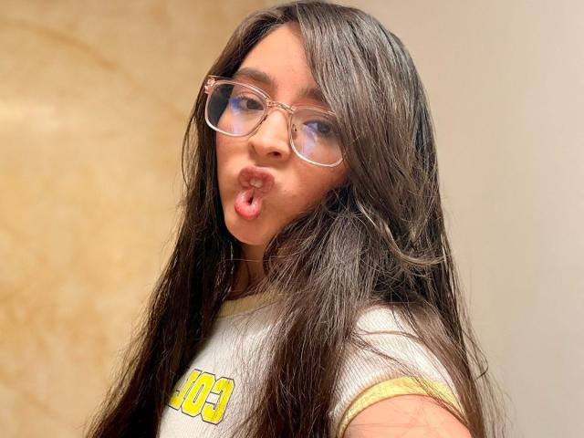 EvaMcAllister with long dark brown hair, wearing clear glasses and a yellow-trimmed white shirt, pouting her pink lips. Her brown eyes are visible behind the glasses.