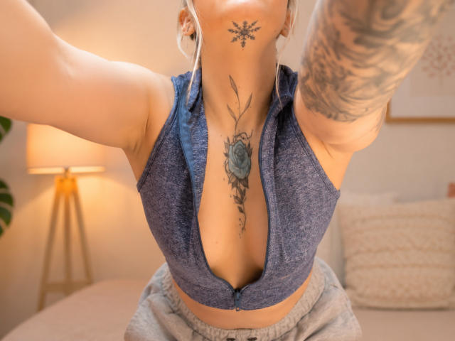 KayleeM with large tits, blue zip-up top, tattoos on throat and arm, blonde hair, earbuds, gray pants, taking a selfie, soft indoor light, cozy room.