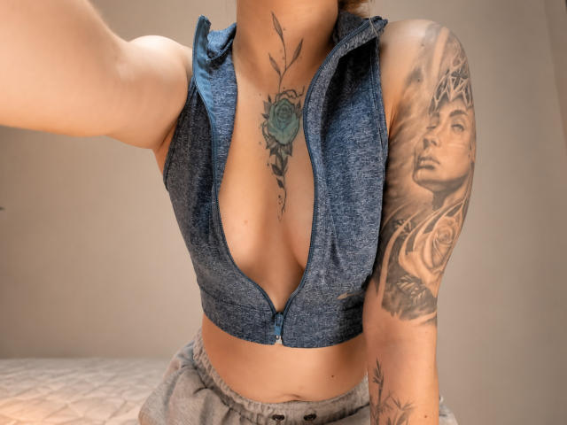 KayleeM with deep cleavage in a blue zip-up top, showing tattoos on chest and arm. Her tits are exposed, gray pants, arm tattoo of a woman's face.