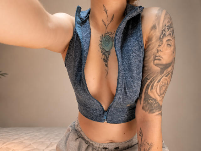 KayleeM with a deep blue zip-up vest, exposing her tits, has tattoos on her chest and arm, and wears gray pants, taking a selfie.