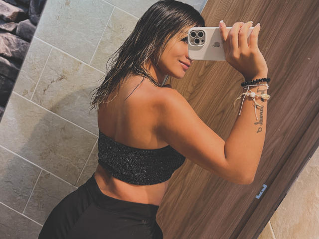 SashaPolet with wet brown hair, wearing a sparkly black crop top and black pants, taking a mirror selfie, showing a tattoo on her right forearm, holding an iPhone.