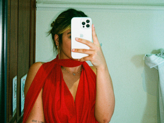 SashaPolet taking a mirror selfie, wearing a red halter dress, holding an iPhone, blonde hair in a messy bun, small tattoo on left arm, necklace, partial view of chest.