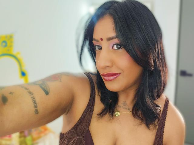 Nahirr with dark hair, pink eyeshadow, red dot on forehead, brown lace top, red lipstick, tattooed arm, gold necklace, smiling, selfie, medium breasts, tan skin, bedroom.