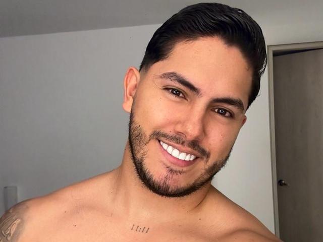 OwenPierce, shirtless, smiling with brown eyes, dark hair, beard, tattoo on chest, and a cute, happy face. His brown eyes and white teeth are prominent.