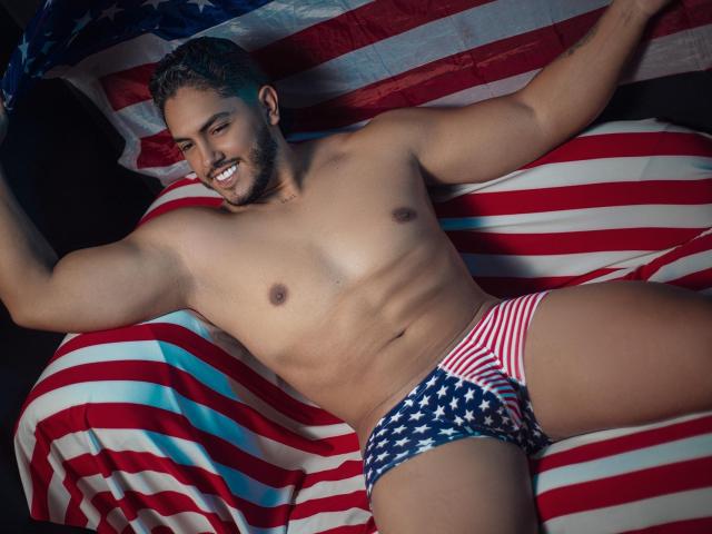 OwenPierce, muscular, tan-skinned, smiling, wearing red/white/blue striped and American flag-patterned briefs, lying on a bed with American flags, arms raised, showing defined abs and chest.