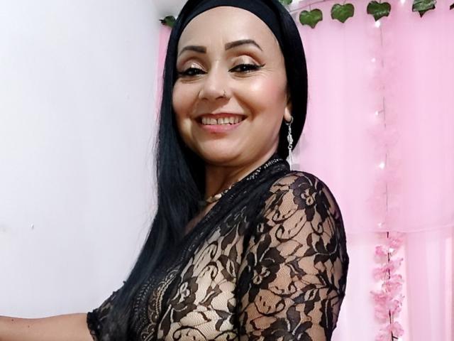 Zaria69 with black lace dress, long black hair, black headscarf, smiling, showing her tits through lace, pink background, green leaves, white wall, and pink string lights.