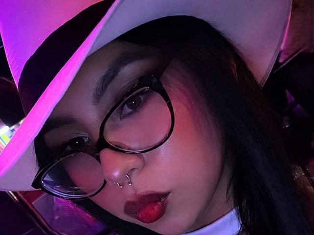 ChloeMoretti with dark hair, glasses, nose ring, and red lipstick, wearing a white hat with a purple glow, looking at the camera. Boobs not visible.