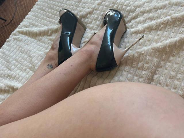MoonLiza's legs with a small tattoo on her right ankle, wearing shiny black high heels with stiletto heels, resting on a textured white blanket.