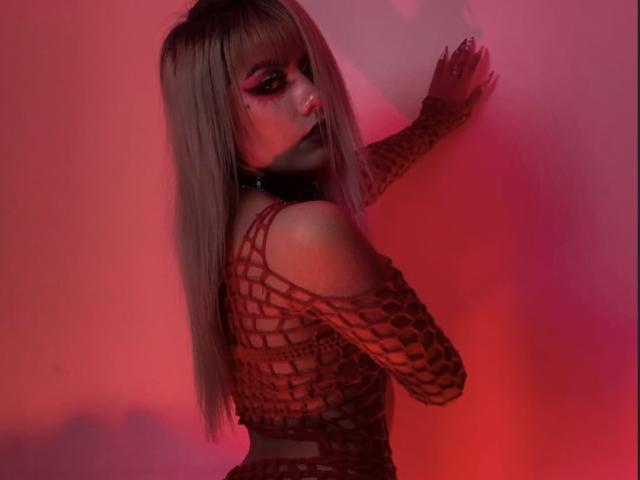 LitenKiitt has long blonde hair, red eyeliner, and wears a red fishnet top, exposing her small tits and a black choker. Red lighting highlights her pale skin.