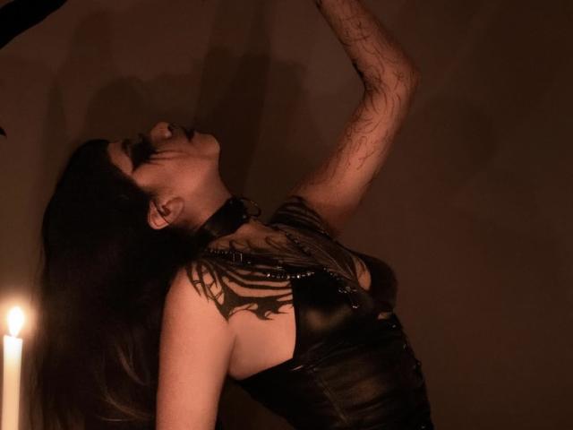 LitenKiitt with dark hair, black leather top, and collar, arms raised, eyes closed, facing upward, candle light in background, tattoos on arm, shadowed, moody, sensual.