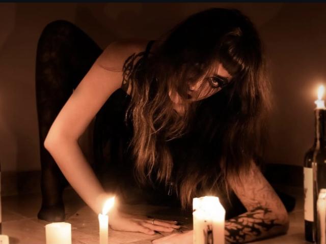 LitenKiitt, crouching, with long brown hair, black lace stockings, and a black top, reaching towards candles, her arms tattooed, in a dimly lit, candle-lit room.