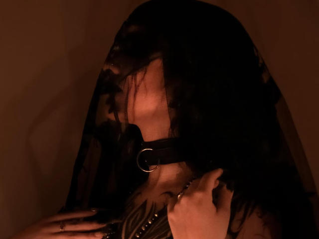 LitenKiitt with black lace veil, wearing a black leather collar with a metal ring, hand on chest, dark brown lighting, semi-transparent lace, visible facial shadow.