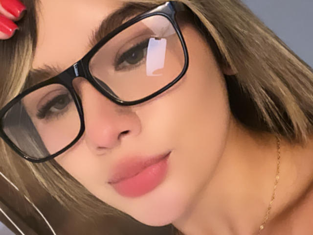 SashaPolet's face with black glasses, pink lips, and blonde hair, holding her red-nail-painted hand on her head. She's wearing a gold necklace.