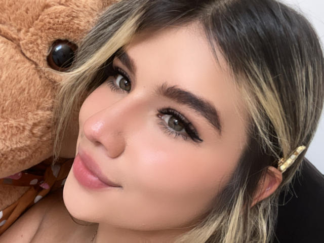 SashaPolet's face with blonde and black hair, brown eyes, and a gold hairpin, showing a slight smile with pink lips, and a fluffy brown teddy bear on the left.