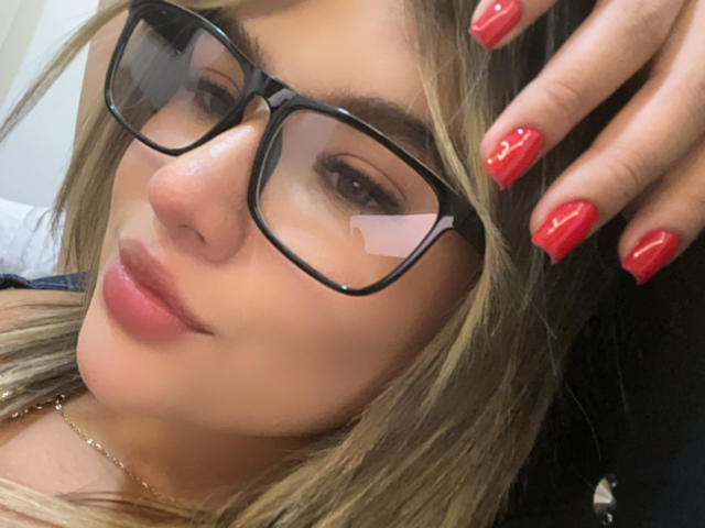 SashaPolet with red nails, glasses, and blonde hair, looking at camera, slightly smiling, wearing a gold necklace, holding her hair.
