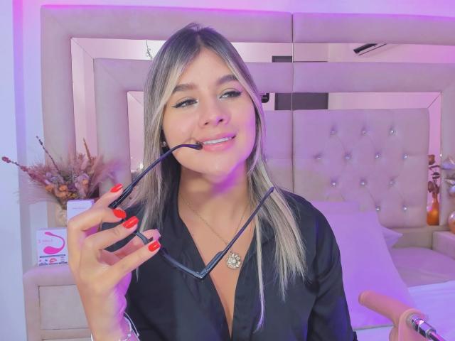 SashaPolet with blonde hair, black robe, holding a black toothbrush in her red-manicured hand, smiling, wearing a necklace with a ring, in a bedroom with purple lighting.
