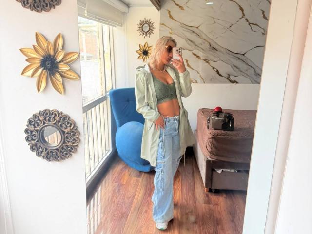 ElenaSaenz with blonde hair, green bra, open shirt, and high-waisted jeans, taking a mirror selfie, showing her tits and ass, in a room with wooden floors.