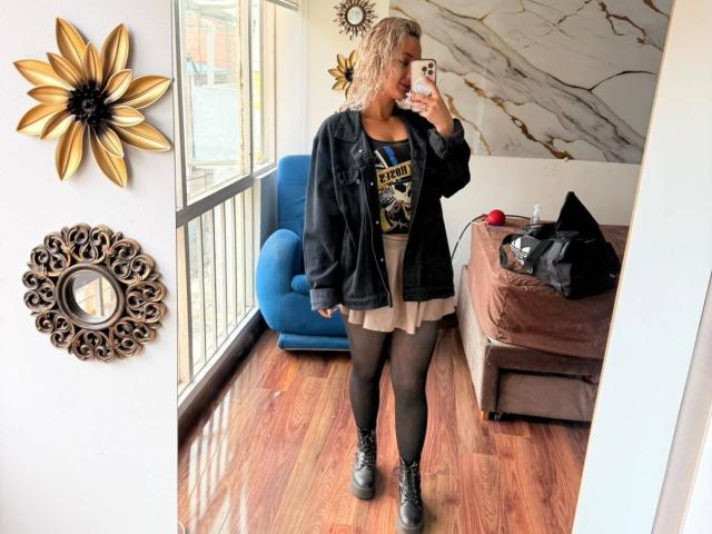 ElenaSaenz stands in a room, wearing a black jacket, graphic tee, beige skirt, black tights, and black boots, holding a phone, with curly blonde hair. Mirror selfie.
