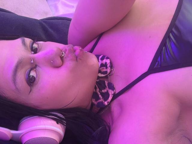 TarahDustin lying down with pink-purple lighting, wearing a leopard print choker, black strappy top, and pink headphones, showing nose piercings and lip piercings, with visible tits.