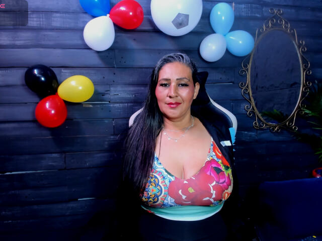 VictoriaKiley with large tits in a colorful, patterned top, long black hair, and a slight smile, sitting in a chair against a dark wooden wall with balloons.