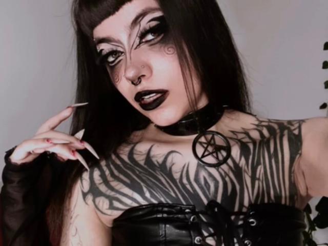 LitenKiitt with goth makeup, black lipstick, tattoos covering her tits, long black hair, black choker with pentagram, black leather corset, and black fingerless gloves, holding a cigarette.
