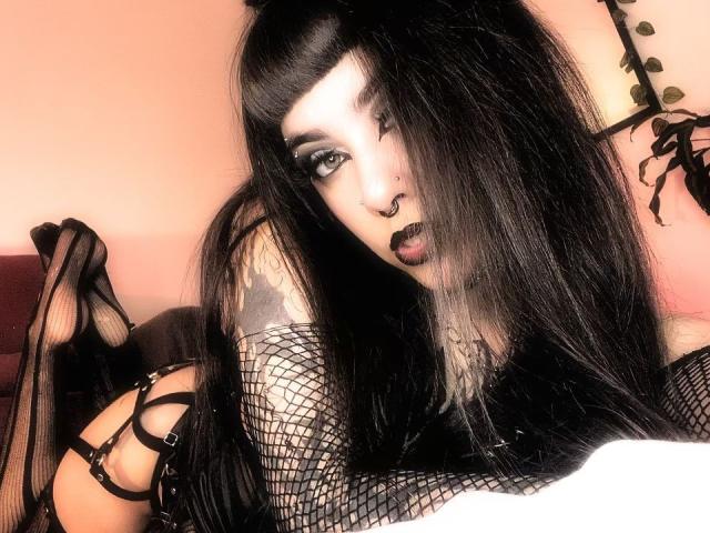LitenKiitt with long black hair, goth makeup, nose ring, dark lipstick, fishnet sleeves, and strappy black lingerie, lying on a bed, showing tattooed arm and high heels.