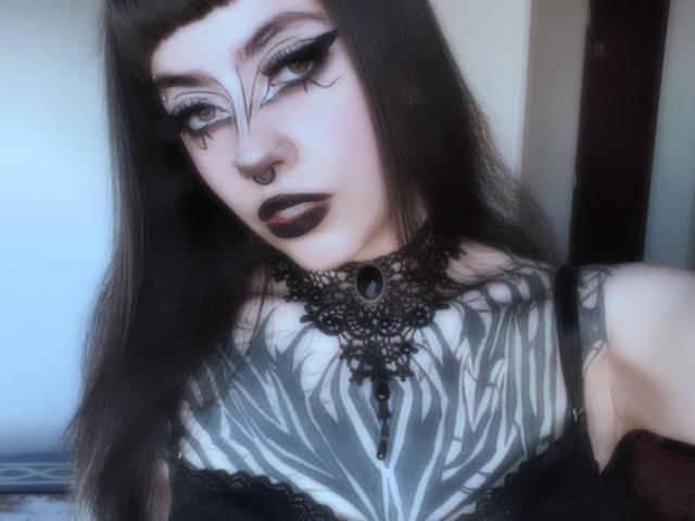 LitenKiitt has dark, long hair, black eyeliner, and black lipstick. She's wearing a black lace choker with a gem and has a zebra-patterned tattoo on her chest.