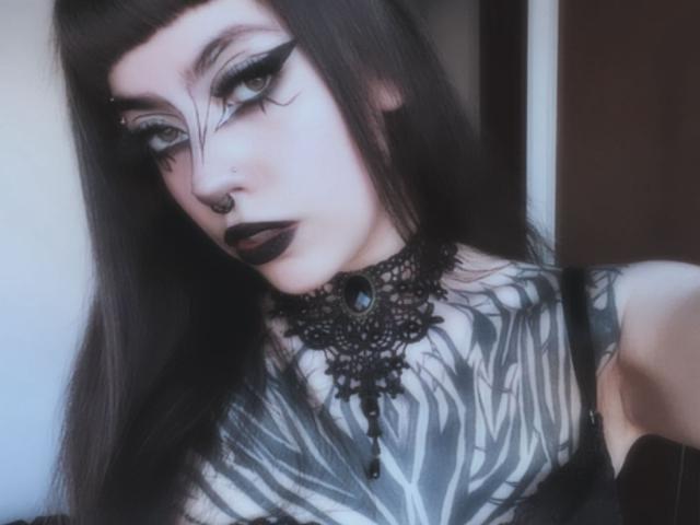 LitenKiitt has goth makeup with thick black eyeliner, dark lipstick, a nose ring, and long black hair. She's tattooed with zebra patterns, wearing a black lace choker.